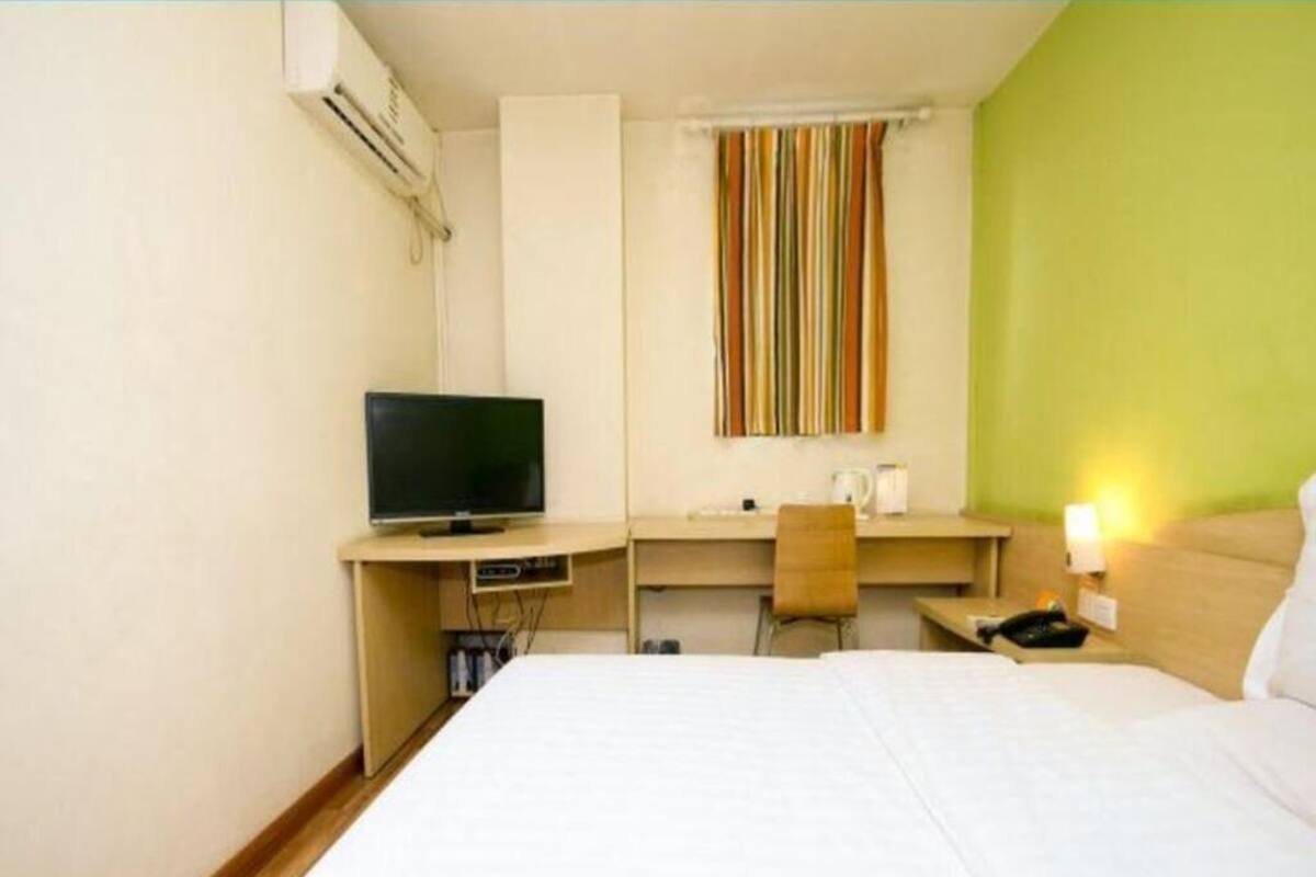 Habitación de hotel con cama, escritorio con televisor, silla, ventana con cortinas a rayas y mesita de noche con lámpara.