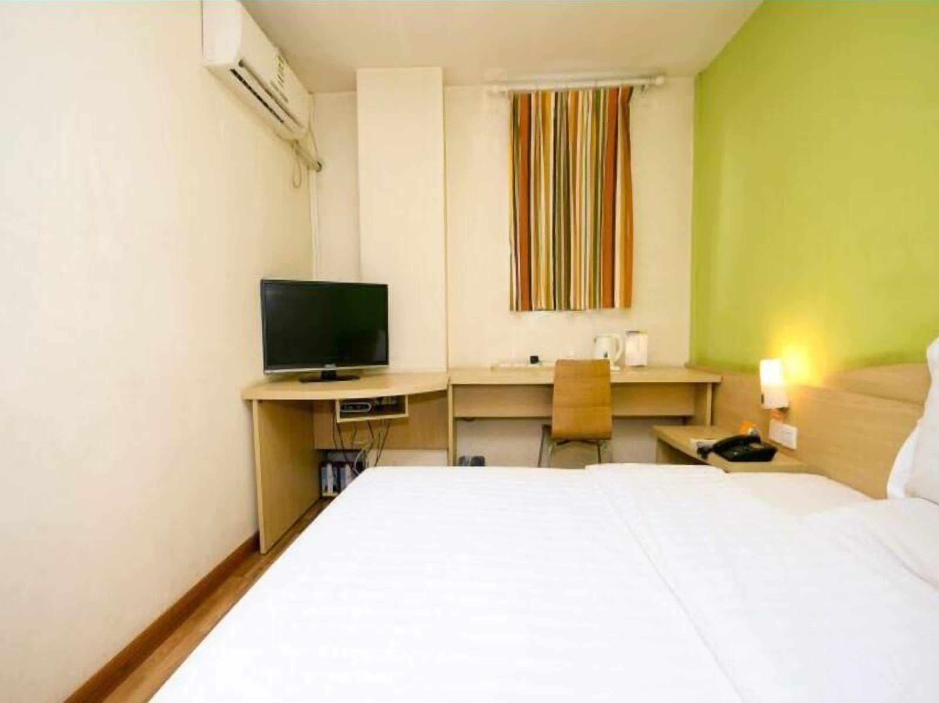 Quarto de hotel com cama, secretária com televisão, cadeira e janela com cortinas listradas.