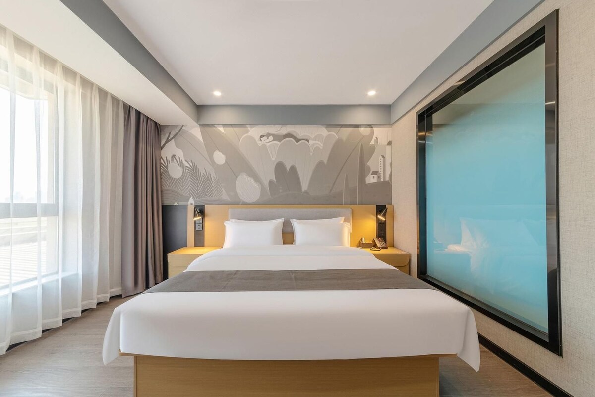 Una habitación de hotel moderna con una cama grande, una mesita de noche, un televisor montado en la pared y vistas de la ciudad a través de una ventana.