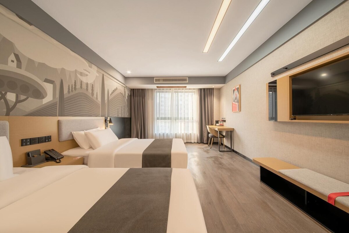 Habitación de hotel con dos camas, un escritorio, un televisor y un ventanal con cortinas.