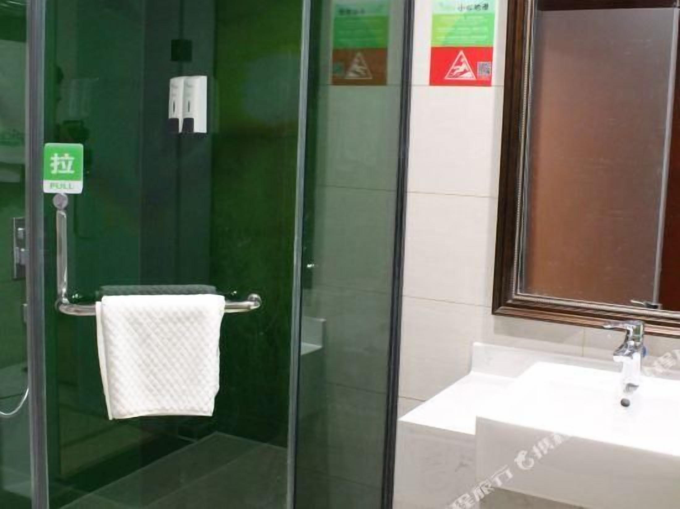Cabina de ducha con paredes de vidrio, una toalla colgada del pomo de la puerta, un lavabo con espejo y una señal de salida verde.