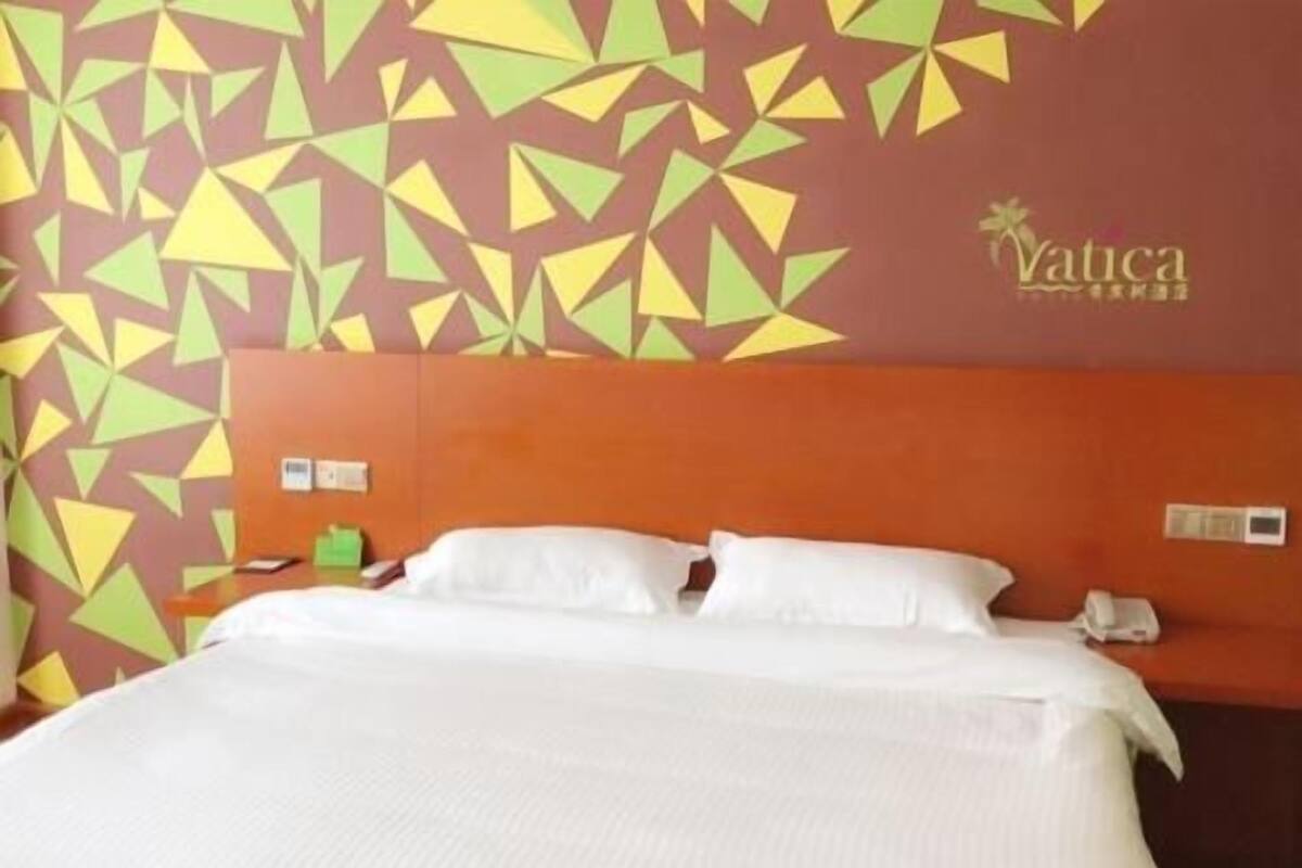 Quarto de hotel com design geométrico na parede, cama com roupa de cama branca e telefone visível na mesa de cabeceira.