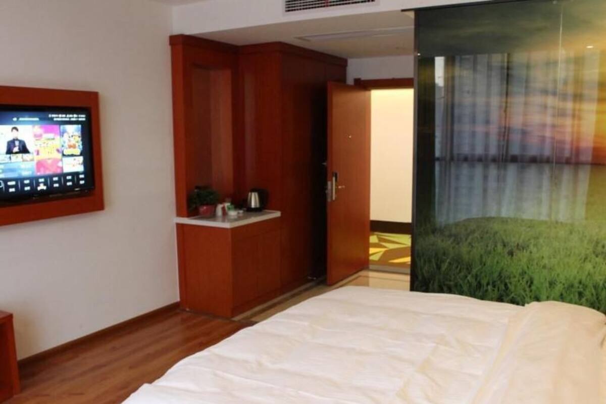 Quarto de hotel com uma cama, uma televisão fixada na parede, um guarda-roupa de madeira e uma porta que dá para outro espaço.