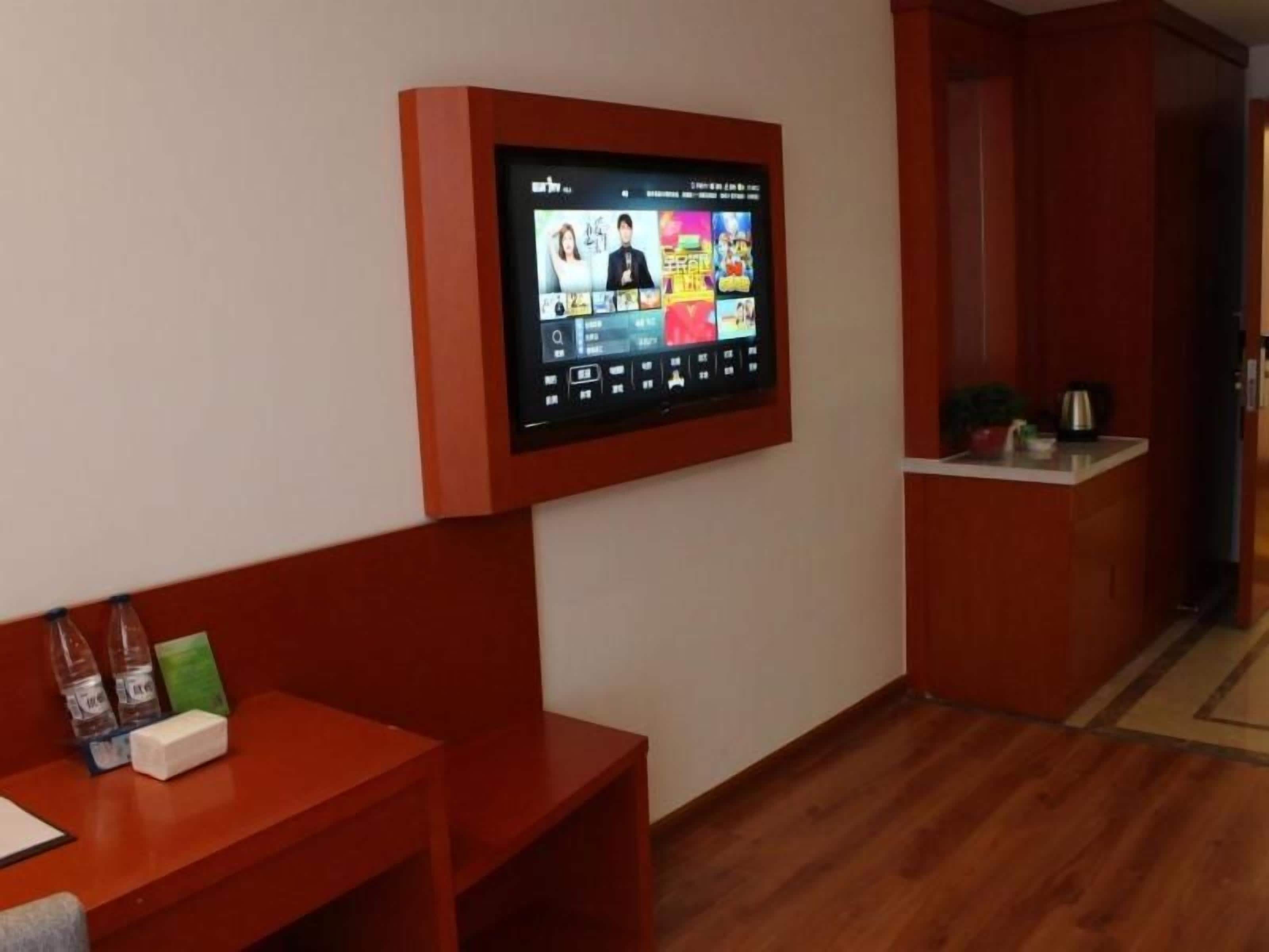 A room with a mounted television displaying a program, a wooden desk with a bottle and a box, and a red bench.
