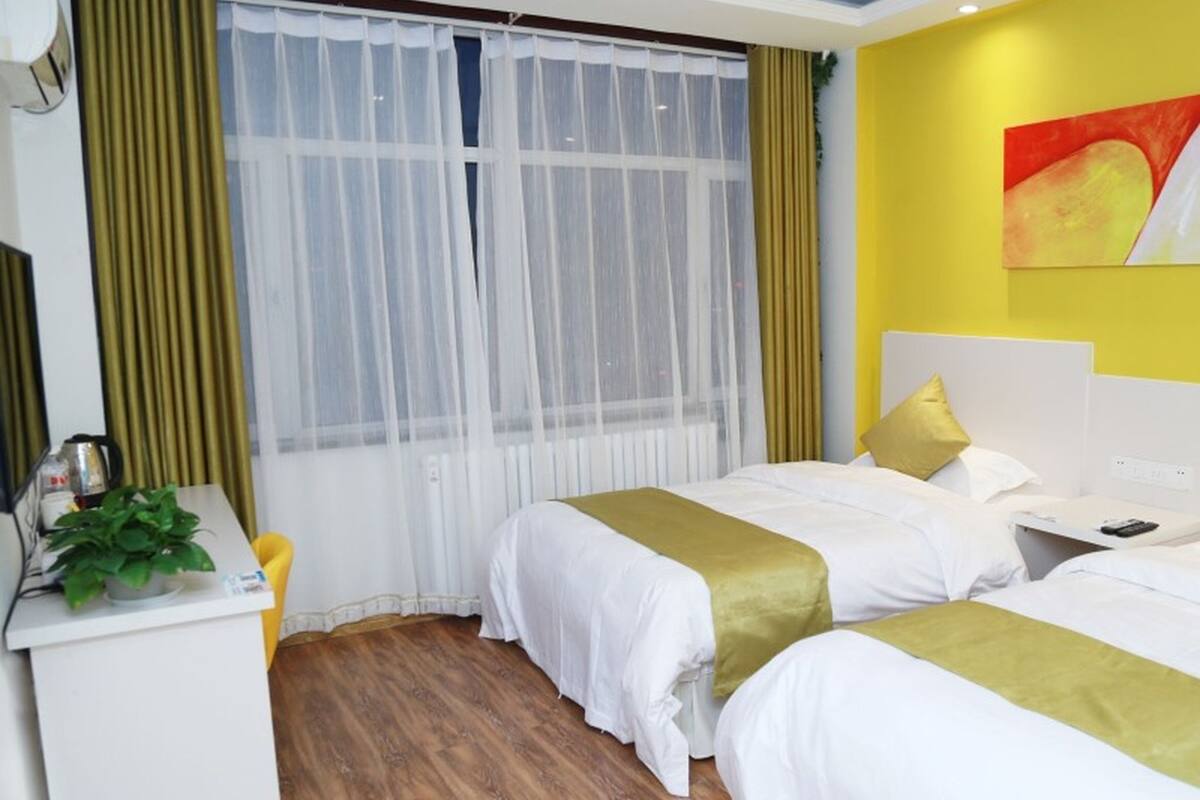 Quarto de hotel com duas camas, uma escrivaninha, uma planta e uma janela com cortinas leves.