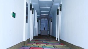 Egy hosszú, keskeny folyosó, színes, mintás szőnyeggel, fehér falakkal és mindkét oldalán ajtókkal.