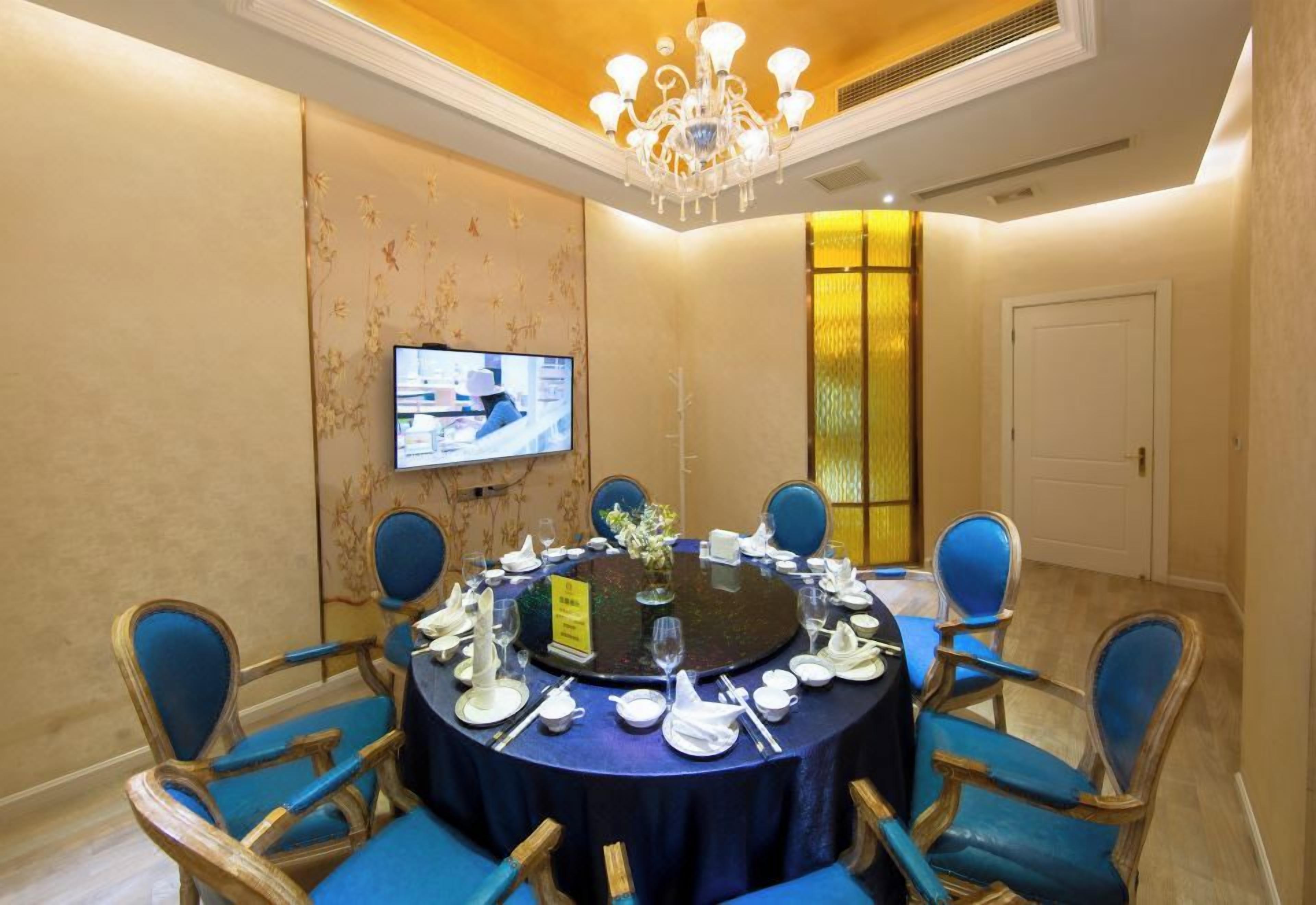 A conference room with a round table, blue chairs, a chandelier, a television on the wall, and a door in the background.