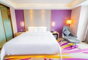 A hotel room with a large bed, a desk, a chair, and a wall-mounted lamp.