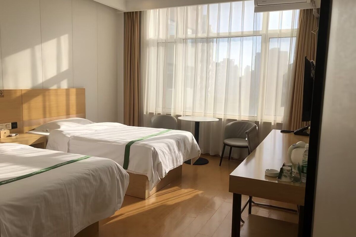 A hotel room with two beds, a desk, a chair, and a large window with sheer curtains.