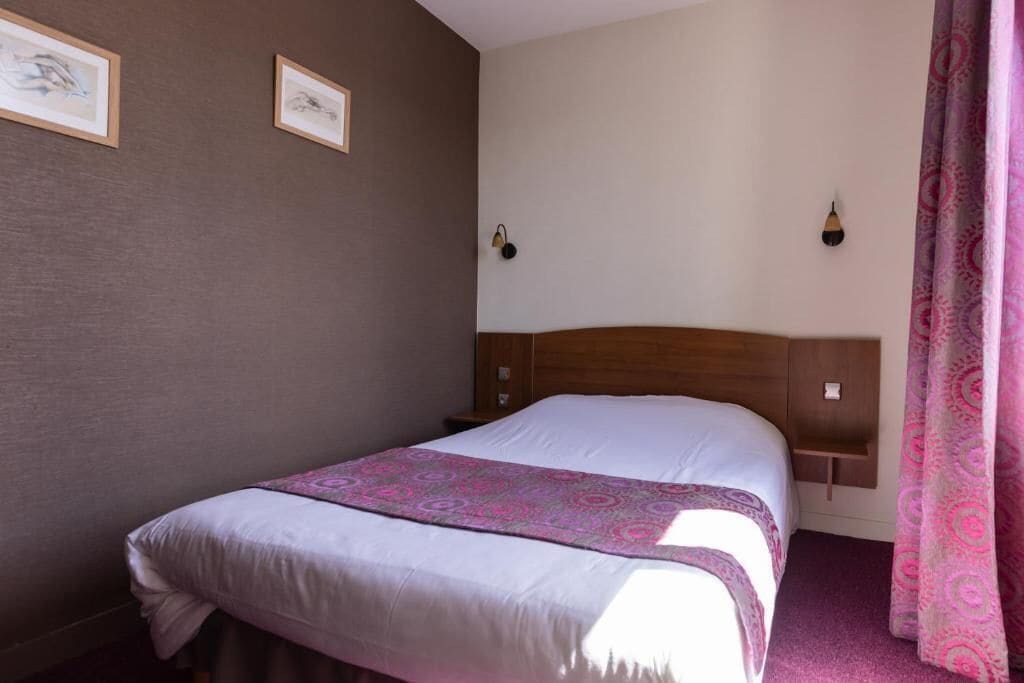 Une chambre d’hôtel avec un lit, une tête de lit, deux appliques murales et une fenêtre avec des rideaux.