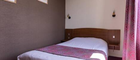 A hotel room with a bed, a headboard, two wall-mounted light fixtures, and a window with curtains.
