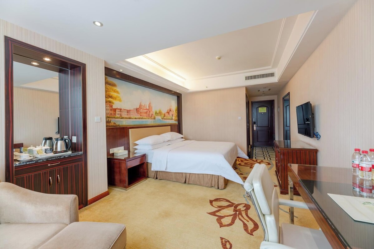 A hotel room with a bed, a desk, a chair, a mirror, a television, and a painting on the wall.