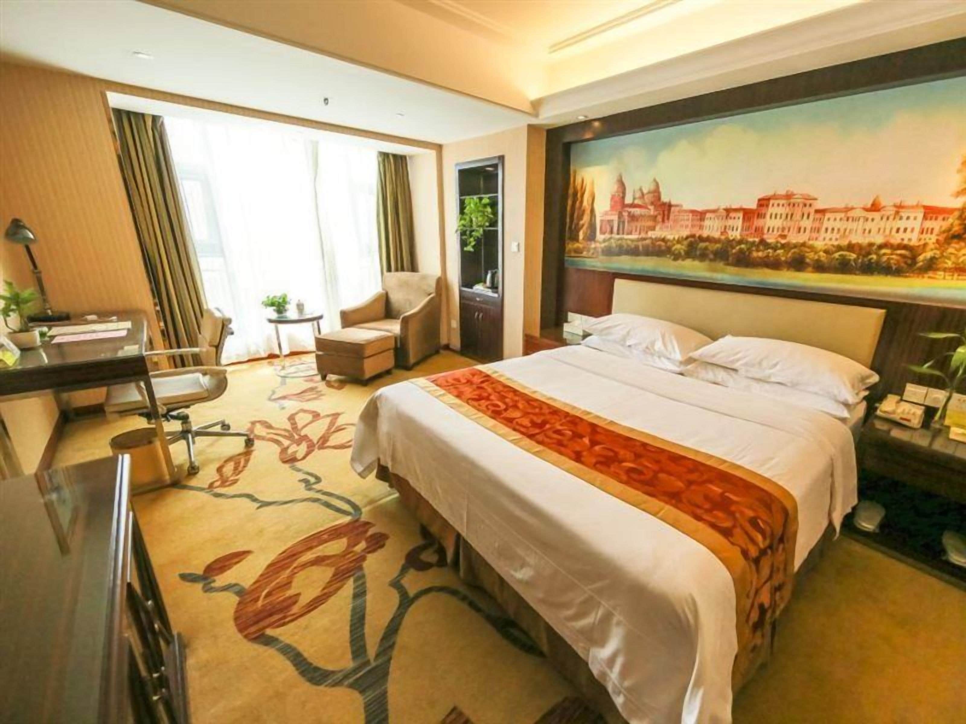 Habitación de hotel con una cama grande, un escritorio, una silla y vistas a un paisaje urbano a través del ventanal.