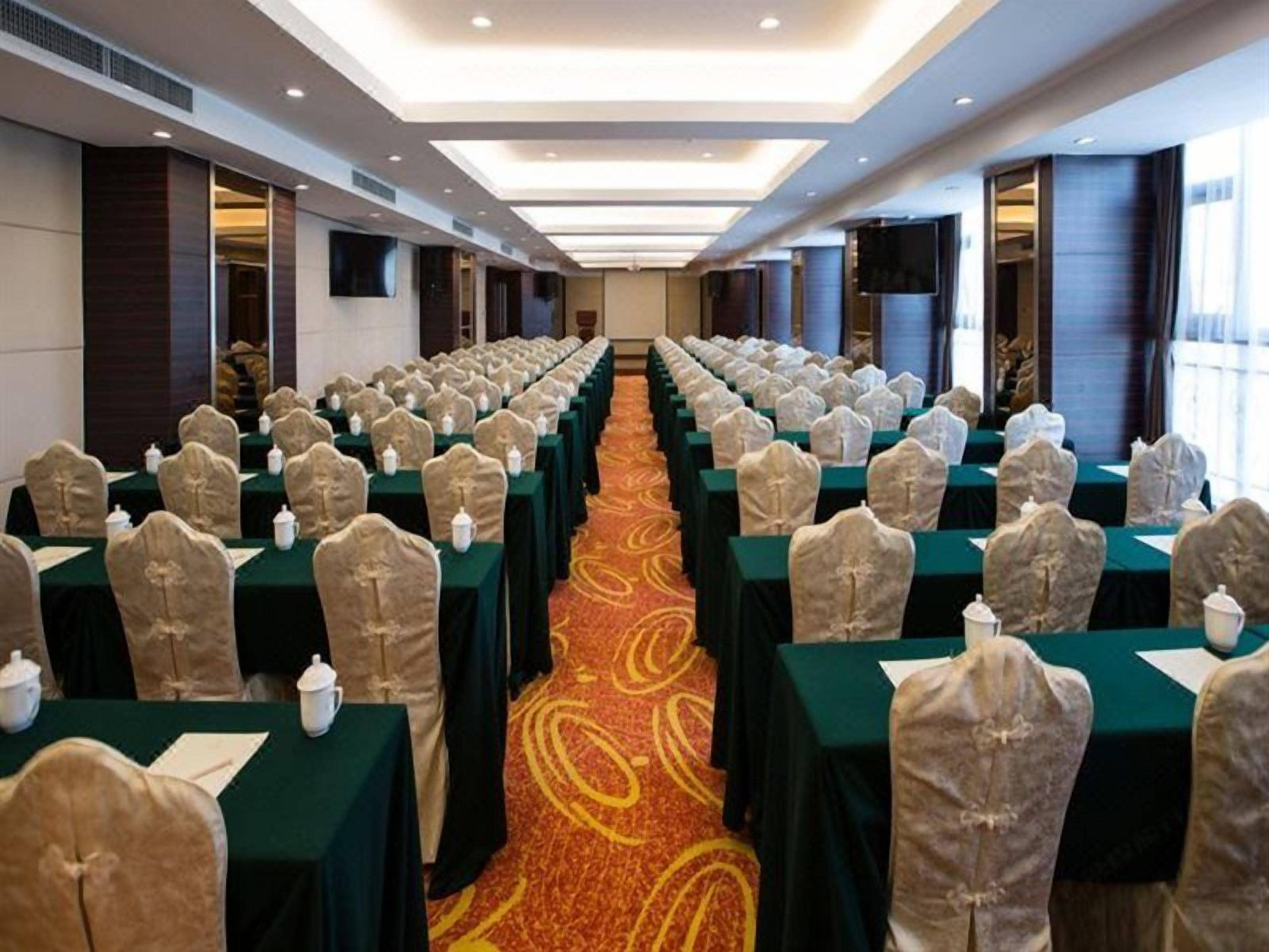 Un salón de conferencias con filas de mesas y sillas, piso alfombrado y una mesa grande al frente.