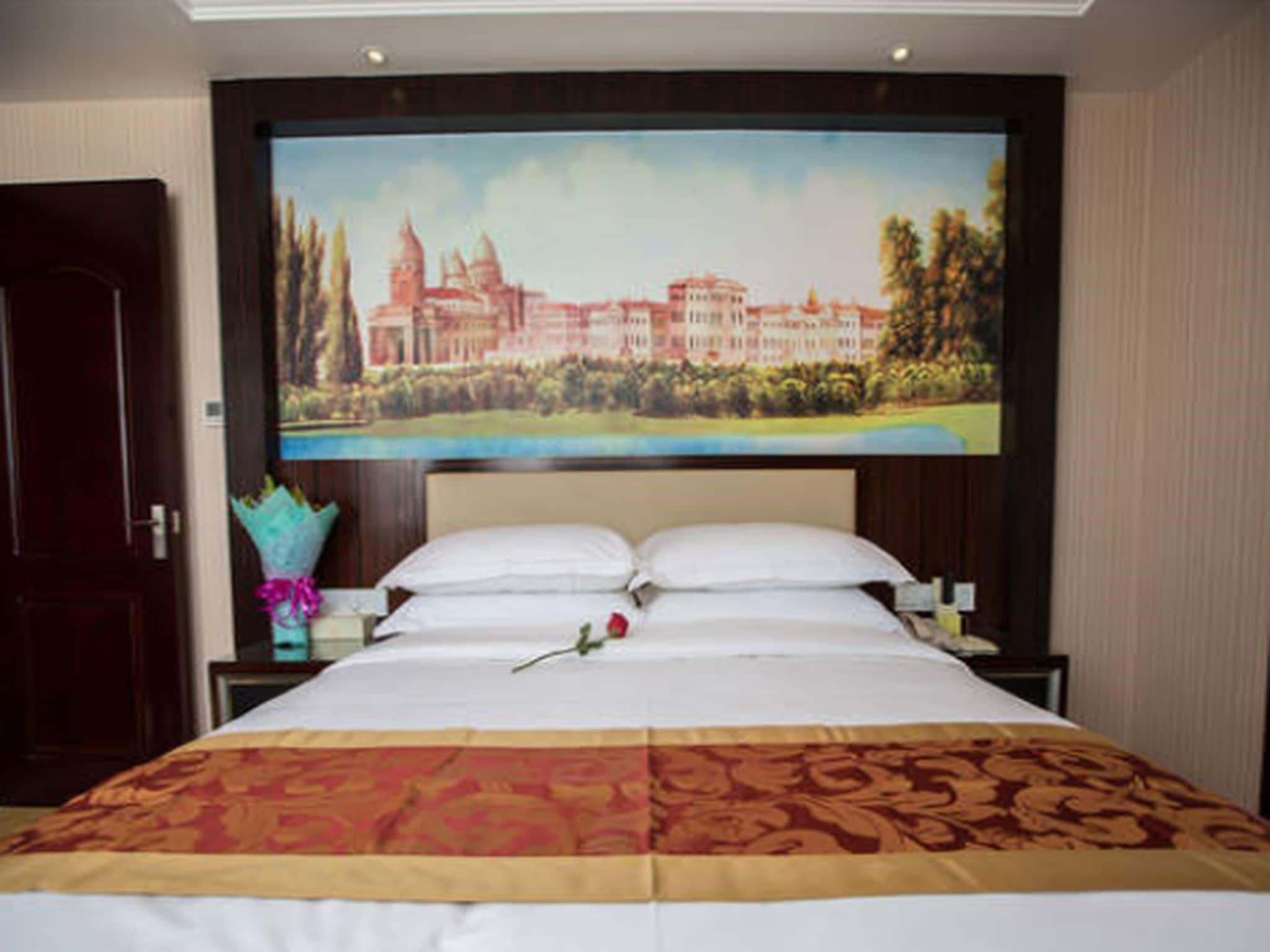 Habitación de hotel con una cama grande, una mesita de noche con un florero y un televisor de pared que muestra un paisaje.