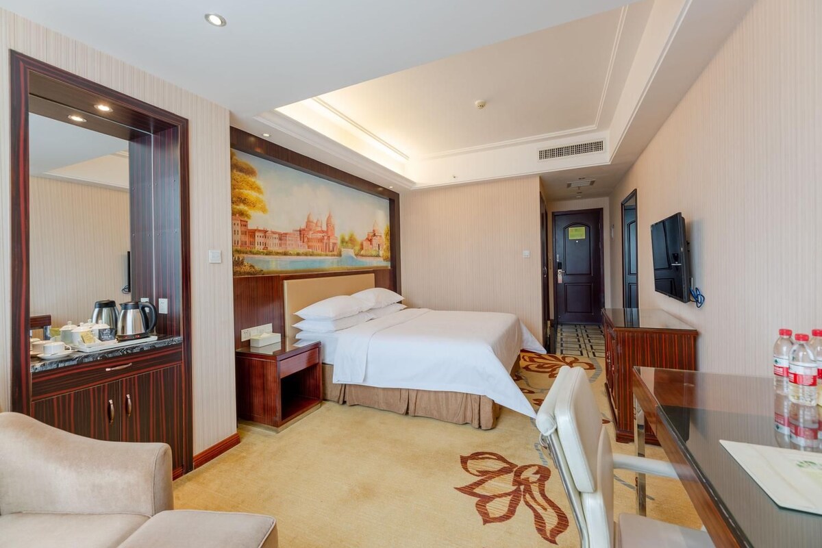 A hotel room with a bed, a desk, a chair, a mirror, a painting, and a television.