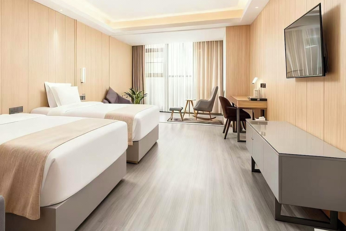 A hotel room with two beds, a desk, a chair, a TV, and a window with curtains.