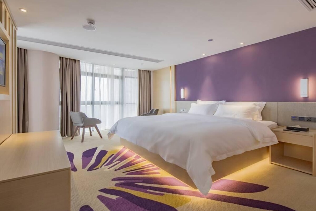 A modern hotel room with a large bed, a desk, a chair, and a view of the outdoors.