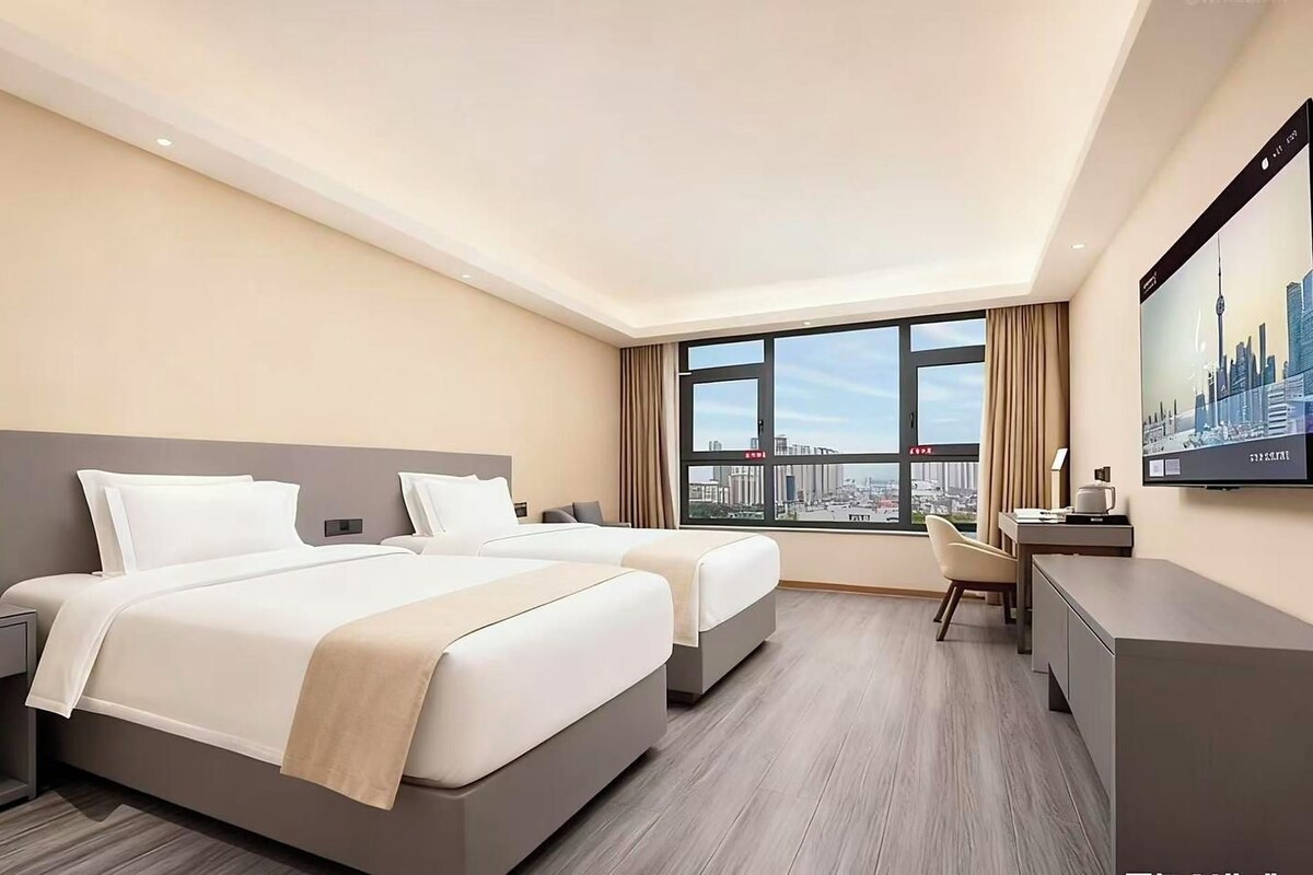 A hotel room with two beds, a desk, a chair, and a large window with a city view.