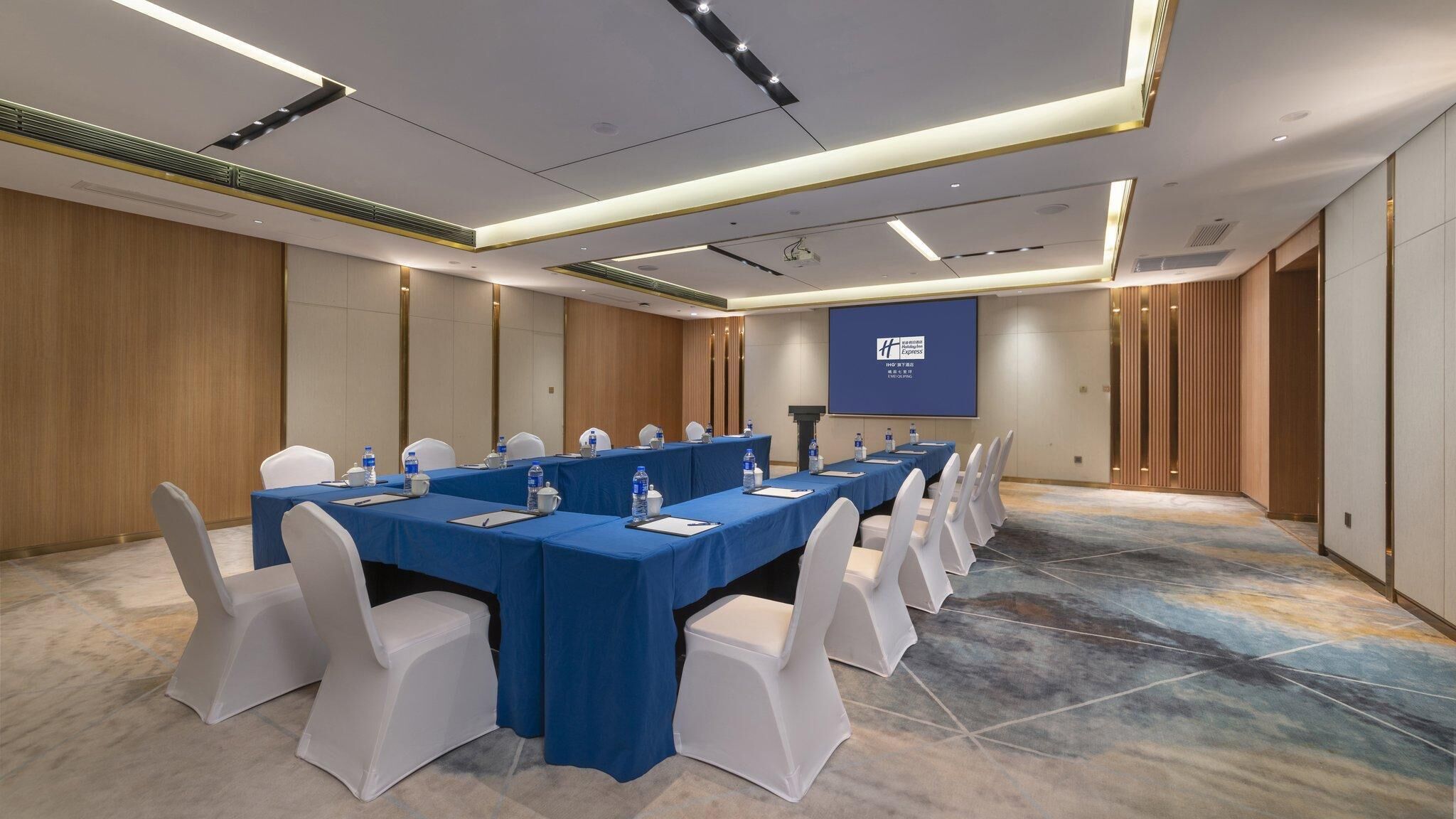 Un salón de conferencias con una mesa larga, sillas blancas y una pantalla para proyector.