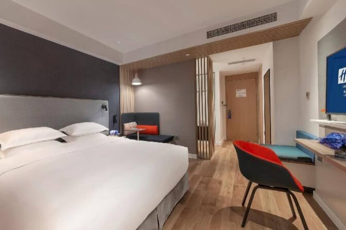 A hotel room with a large bed, a desk with a chair, a TV, and a door leading to another room.