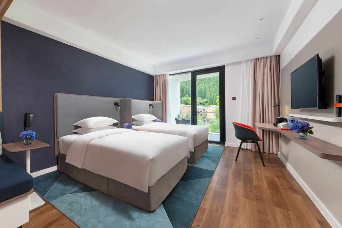 A modern hotel room with two beds, a sofa, a desk, a TV, and a balcony with a view.