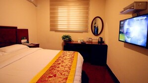 A hotel room with a bed, a desk, a television, a window with blinds, and an air conditioning unit.