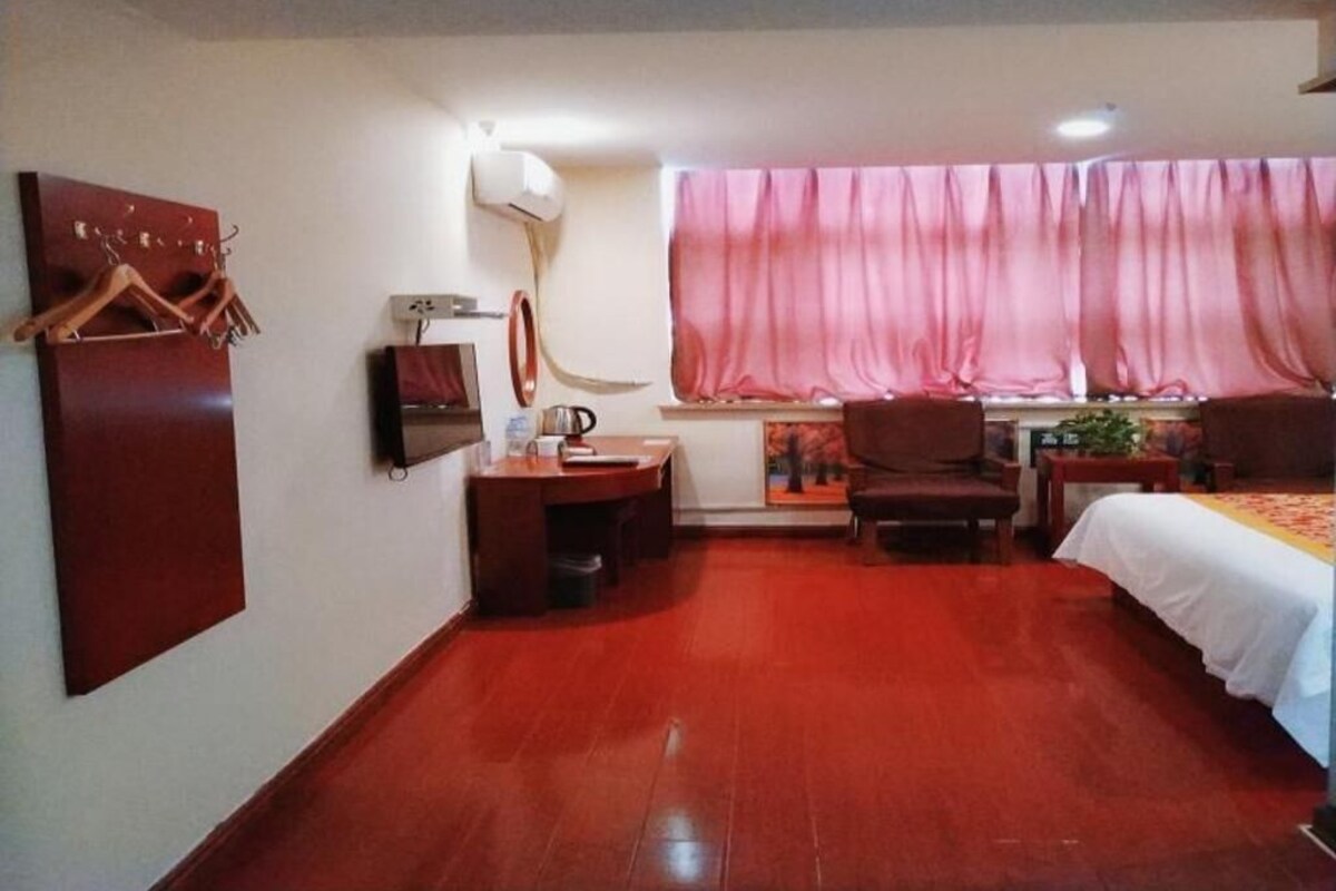A hotel room with a wooden floor, a bed with a white bedspread, a desk with a lamp, a chair, a TV, and a window with pink curtains.