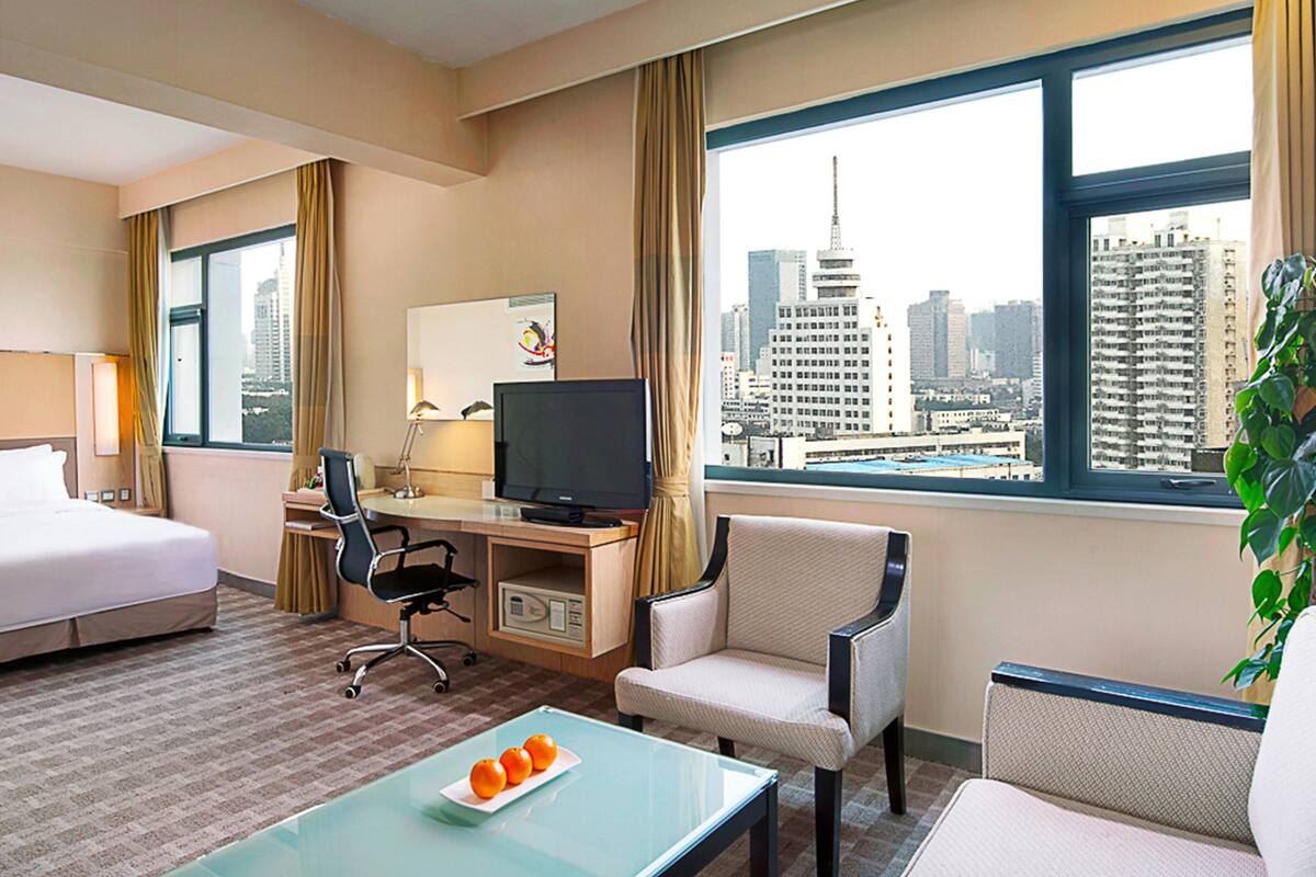 A hotel room with a bed, desk, chair, TV, and a view of a cityscape.