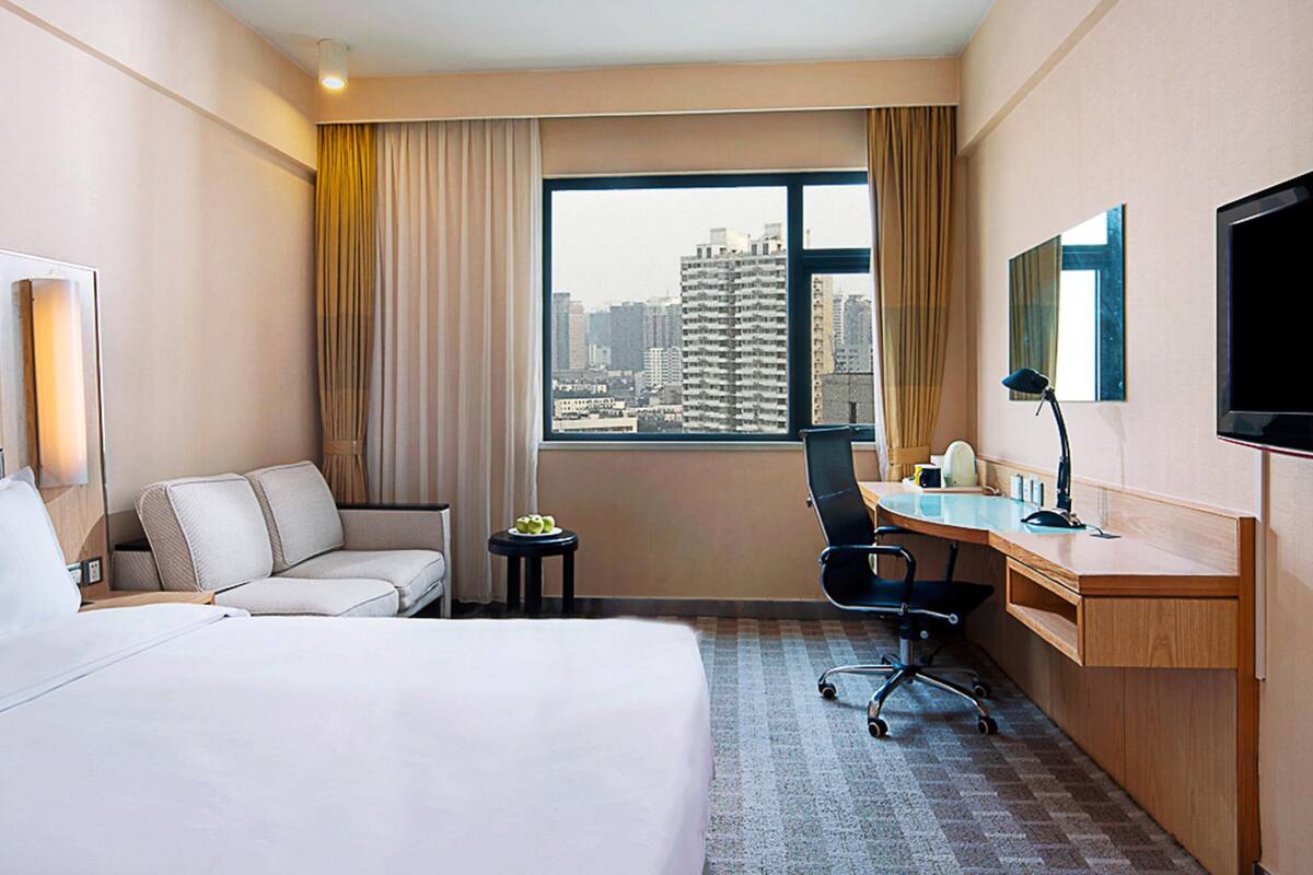 A hotel room with a bed, a desk, a chair, a small table with apples, a TV, and a window with a city view.