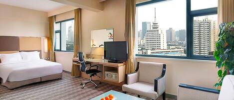 A hotel room with a bed, desk, chair, TV, and a view of a cityscape.