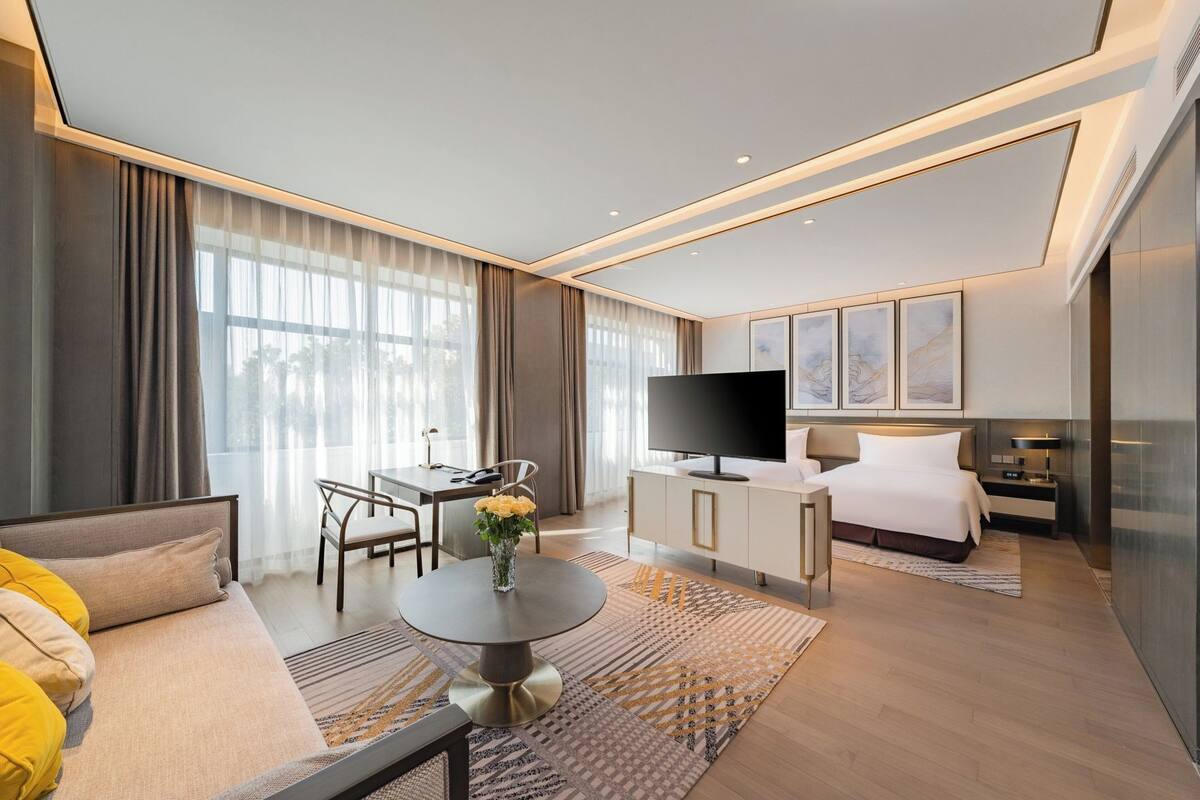 A modern hotel room with a sofa, a dining table with chairs, a TV, a bed, and artwork on the walls.
