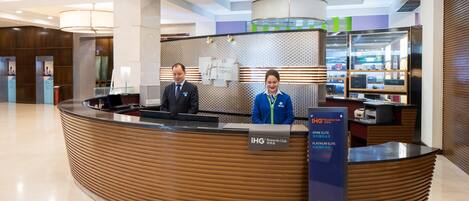 A reception desk with two staff members, a sign for the IHG Rewards Club, and a display of hotel amenities.