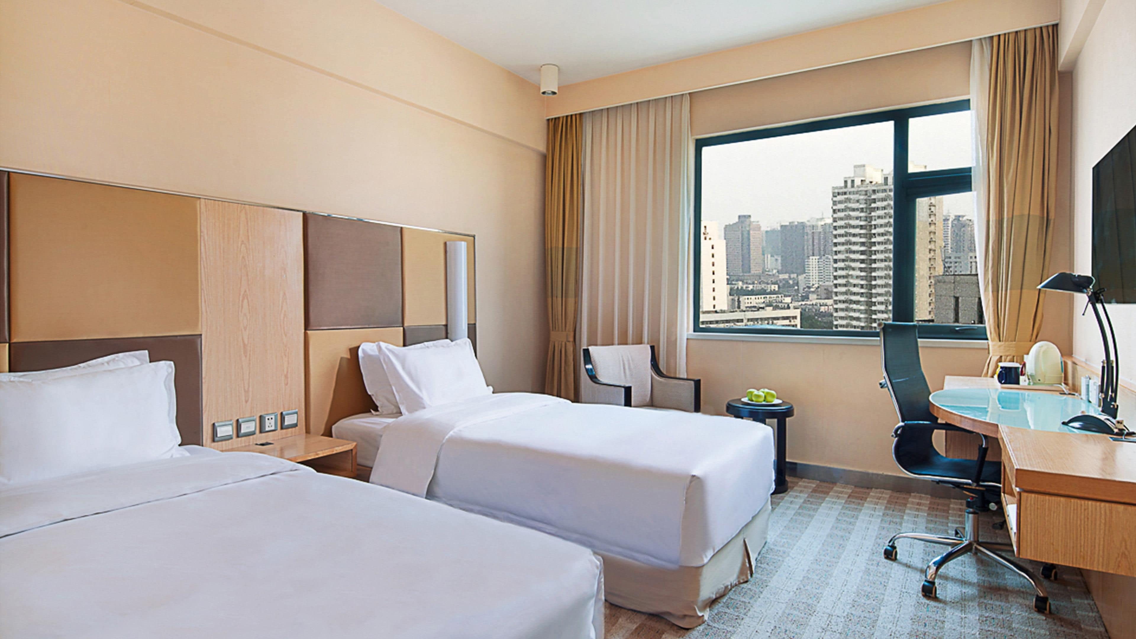 A hotel room with two beds, a desk, a chair, a lamp, and a view of a cityscape through a large window.
