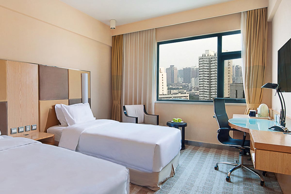 A hotel room with two beds, a desk, a chair, a lamp, and a view of a cityscape through a large window.