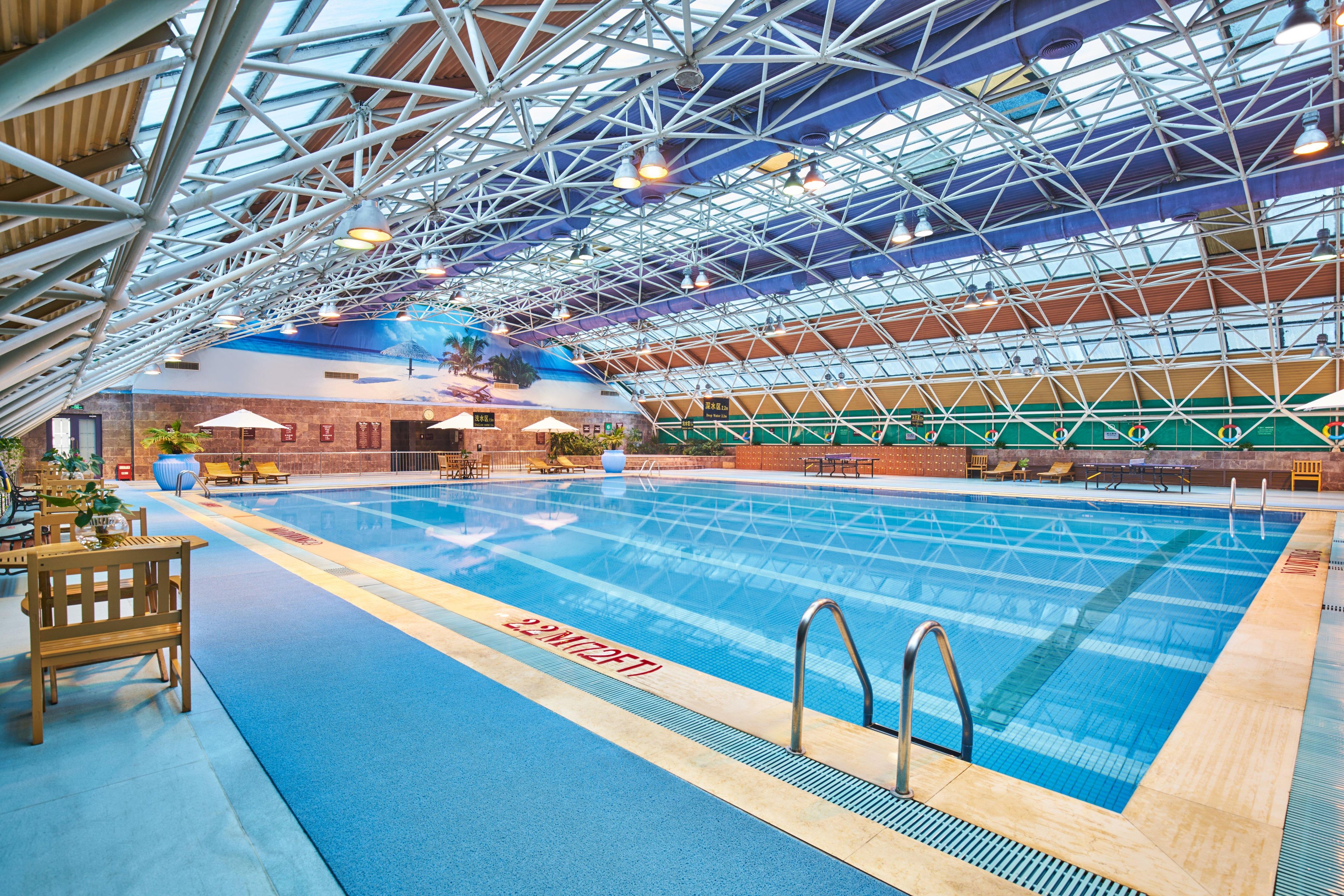 An indoor swimming pool with a high ceiling and metal beams, surrounded by lounge chairs and a blue floor.