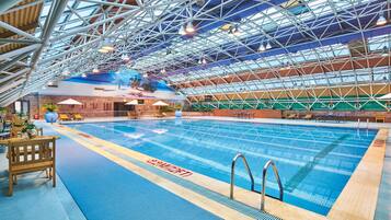 An indoor swimming pool with a high ceiling and metal beams, surrounded by lounge chairs and a blue floor.