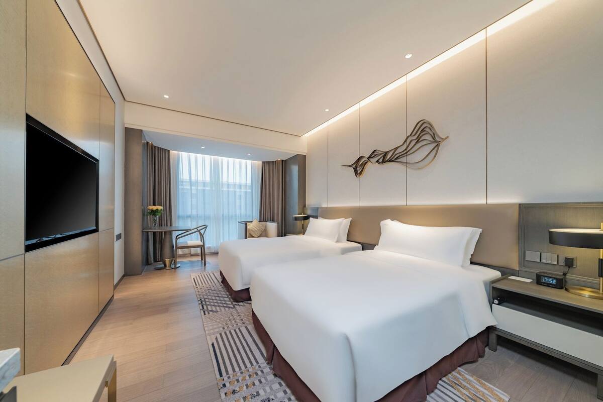 A modern hotel room with two beds, a flat-screen TV, a desk, and a large window.