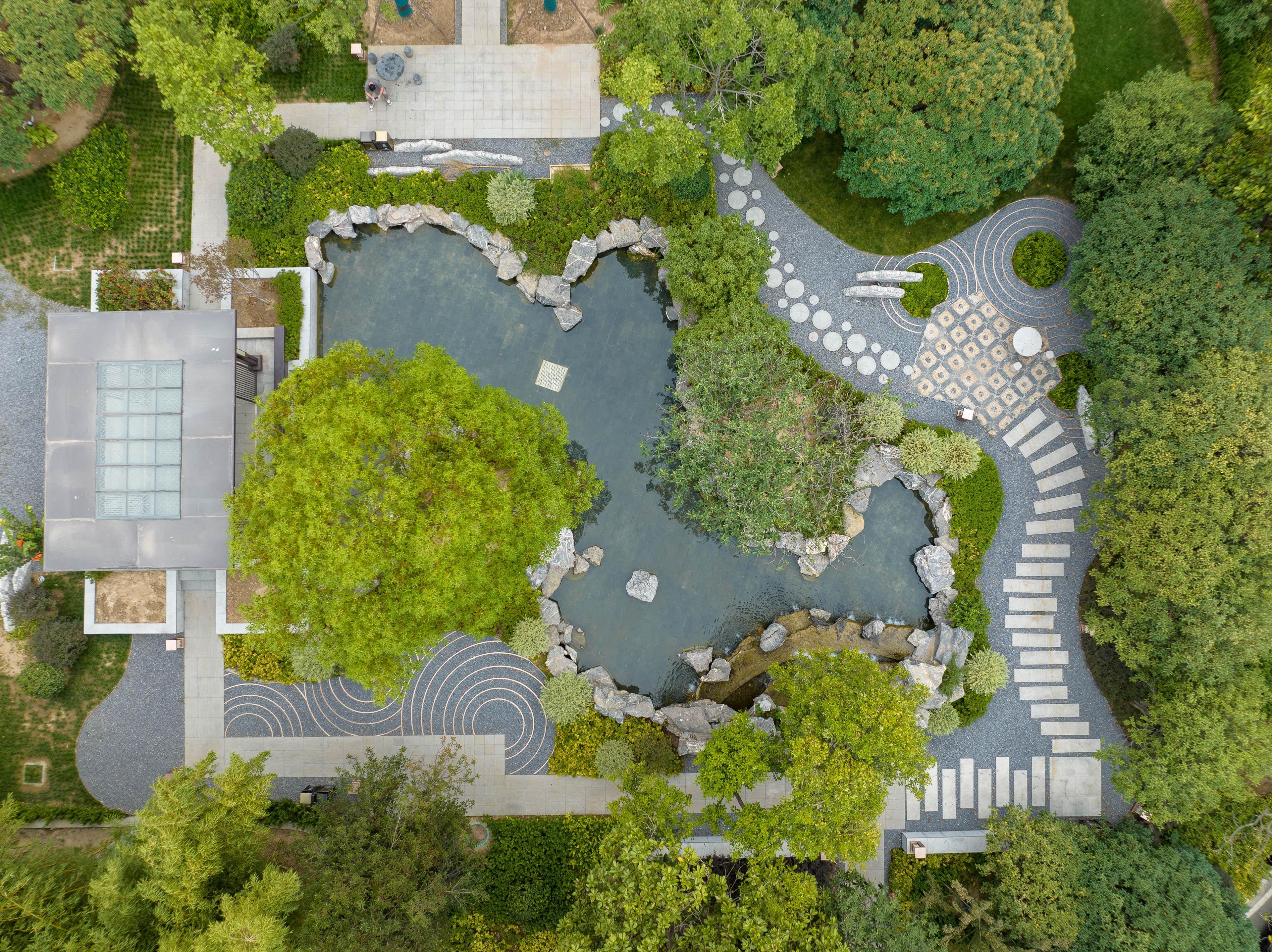 An aerial view of a landscaped area with a central pond, stone pathways, and surrounding greenery.