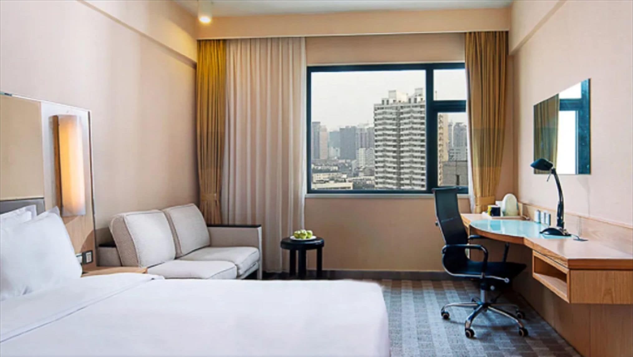 A hotel room with a bed, a desk, a chair, a small table with a plant, and a view of a cityscape through a large window.