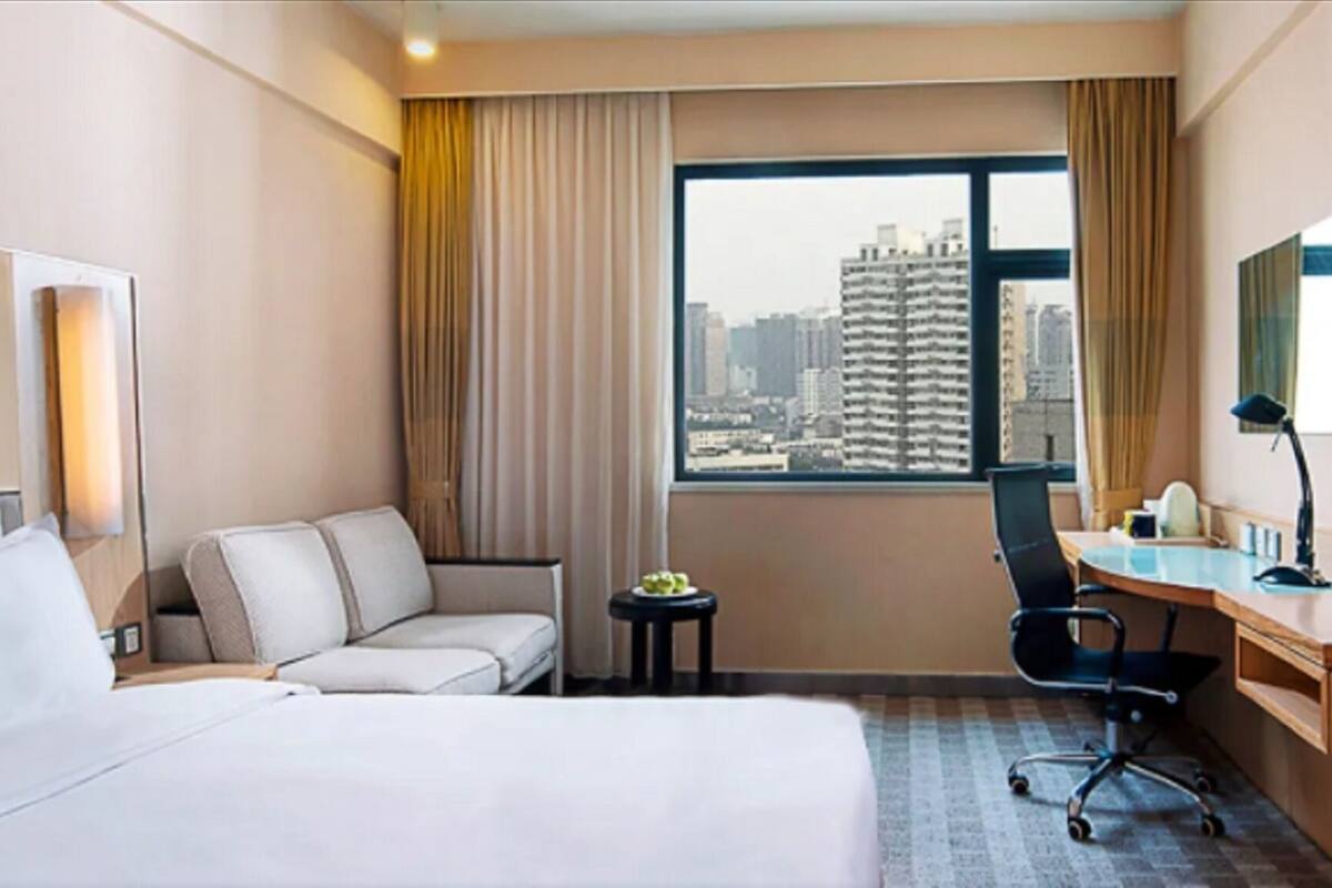 A hotel room with a bed, a desk, a chair, a small table with a plant, and a view of a cityscape through a large window.