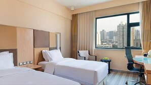 Hotellihuone, jossa on kaksi sänkyä, työpöytä, tuoli, pieni pöytä hedelmillä ja suuret ikkunat, joista avautuu näkymä kaupungin ylle.