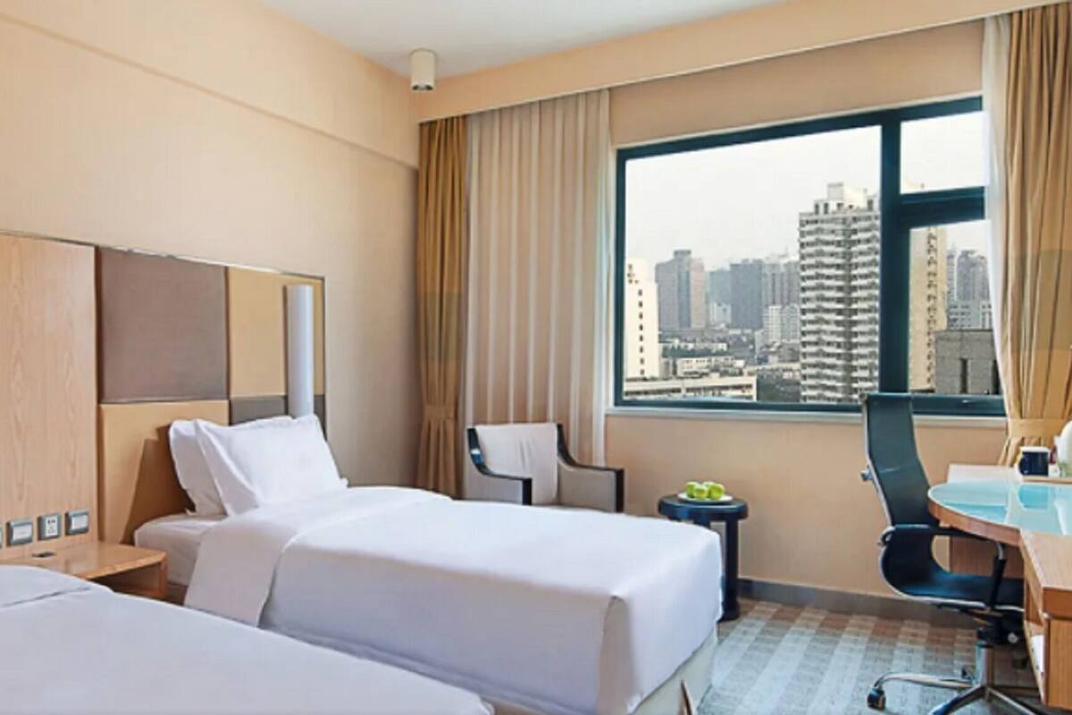 A hotel room with two beds, a desk, a chair, a small table with fruits, and a view of a cityscape through large windows.