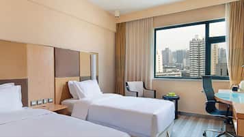 Quarto de hotel com duas camas, uma escrivaninha, uma cadeira, uma mesinha com frutas e vista para a cidade através de grandes janelas.