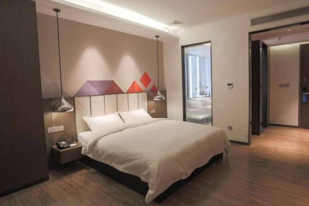 Quarto de hotel moderno com uma cama grande, luminárias, mesa de cabeceira e vista da cidade por uma janela ampla.