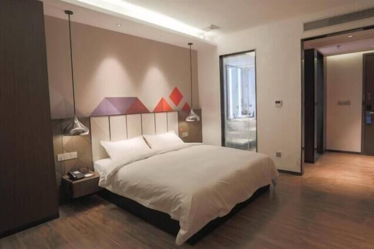 A modern hotel room with a large bed, bedside lamps, a nightstand, and a view of the city through a large window.