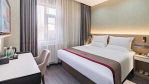 A hotel room with a bed, a desk, a chair, a window with curtains, and a wall-mounted nightstand. - Shankee Hotel (Shunyi)