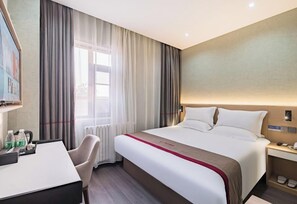 Ein Hotelzimmer mit Bett, Schreibtisch, Stuhl, einem Fenster mit Vorhängen und einem an der Wand befestigten Nachttisch.