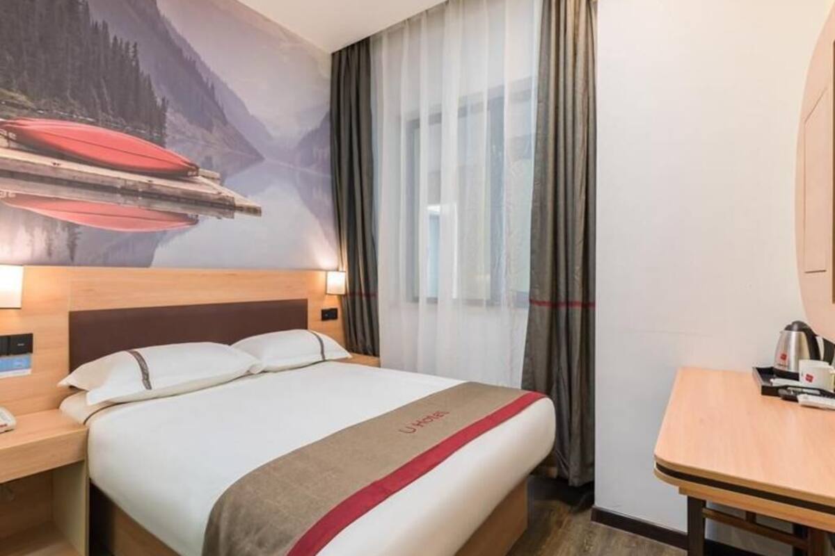 Una habitación de hotel con cama, escritorio, silla, mesita de noche, lámpara y un cuadro de un paisaje montañoso.