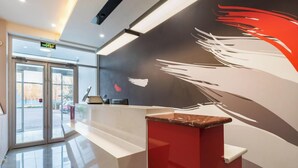 A reception area with a modern design, featuring a reception desk, a glass door entrance, and a wall with abstract art. - Shankee Hotel (Shunyi)
